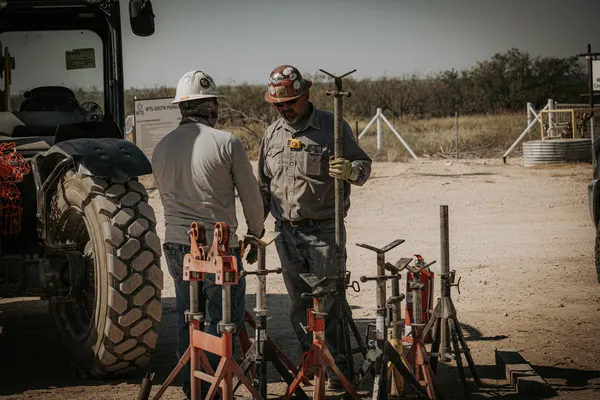 Two construction workers discuss equipment near a tractor and a selection of jacks on a dusty worksite.