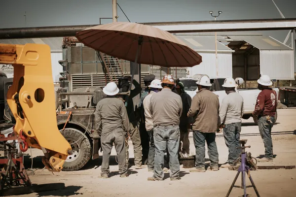 A group of workers in hard hats gathers under an umbrella on a construction site, discussing tasks near machinery.