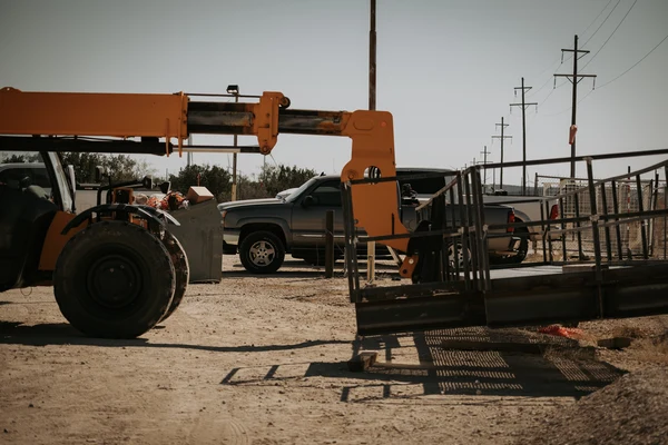 A yellow telehandler loads material while a gray pickup truck is parked nearby on a dusty construction site. Power lines stretch overhead.