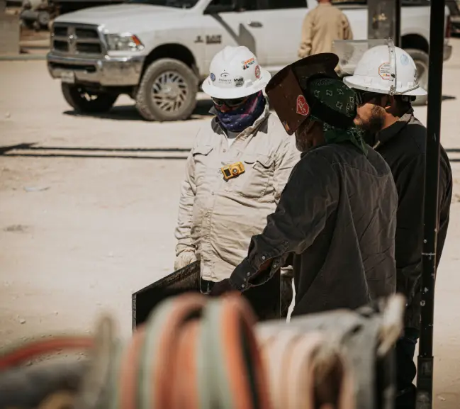 about img 11 Three construction workers in hard hats and safety gear discuss over equipment, with a pickup truck parked nearby on a job site.