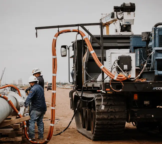 about page img 1 Two workers in hard hats operate machinery on a construction site, with hoses connected to a large industrial truck.