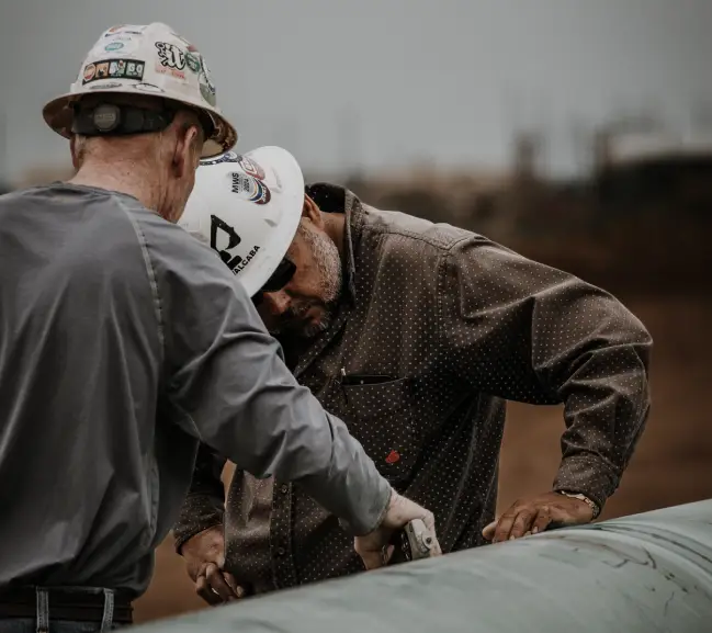 Two workers in hard hats examine a large pipe, focused on their task against a muted industrial background.
