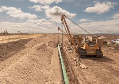 Two yellow bulldozers work to install a green pipe in a dug trench on a construction site under a cloudy sky.