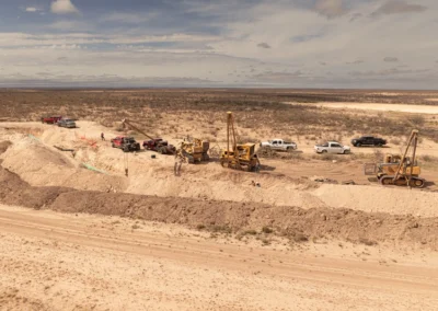 Construction equipment and workers operate in a desert landscape, surrounded by trucks and machinery, with a cloudy sky above.