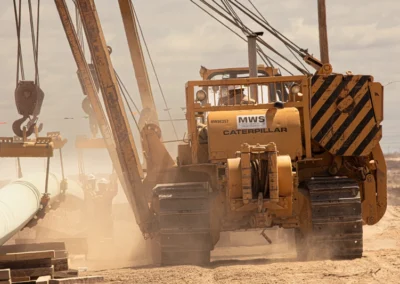 A yellow Caterpillar machinery lifts a large pipe on a dusty construction site under a cloudy sky.