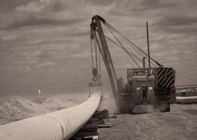 A large caterpillar crane hoists a pipeline section amid dusty terrain, with power lines and a distant flame in the background.