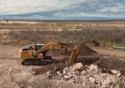 A CAT excavator digging into earth, with large piles of dirt and rocks in an expansive, open landscape under a cloudy sky.