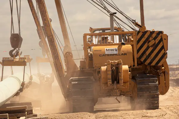 Caterpillar machine at a construction site, lifting a large pipe amidst dust and sunny skies.